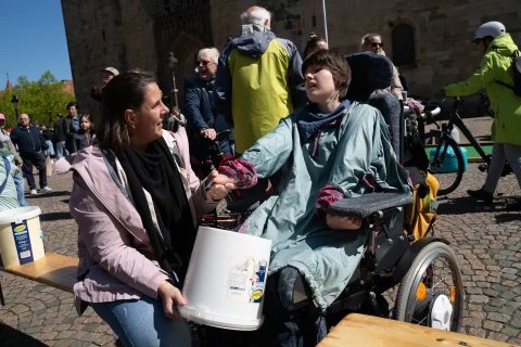 Zwei Personen im Au&szlig;enbereich auf einem gepflasterten Platz. Eine Person sitzt in einem Rollstuhl und tr&auml;gt eine blaue Jacke, die andere kniet links daneben und tr&auml;gt eine rosafarbene Jacke und eine schwarze Schal. Beide halten gemeinsam einen wei&szlig;en Eimer mit einer Zeichnung darauf. Im Hintergrund sind mehrere Menschen sowie eine sandfarbene Geb&auml;udefassade sichtbar.