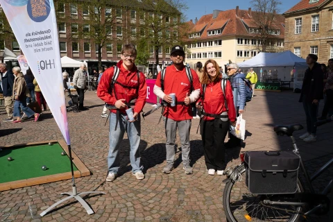 Vier Personen in roten Jacken mit Rucks&auml;cken stehen auf einem gepflasterten Platz. Links ein Banner, im Vordergrund ein Fahrrad, rechts Passanten. Im Hintergrund Marktst&auml;nde, B&auml;ume und Geb&auml;ude mit roten D&auml;chern.