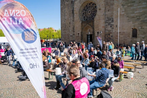 Menschen sitzen auf langen Holz-B&auml;nken auf einem gepflasterten Platz vor einer gro&szlig;en, steinernen Kirchenfassade mit Rosettenfenster. Links steht eine bunte Fahne mit Text &bdquo;OSNA F&Uuml;R ALLE&ldquo; und QR-Code. Im Hintergrund weitere Menschen, St&auml;nde und gr&uuml;ne B&auml;ume unter blauem Himmel.