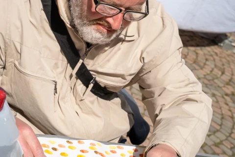 Person in heller, beige Jacke sitzt an einem Tisch, malt mit einem Pinsel auf ein Blatt mit vielen bunten Punkten. Rechts unten am Tisch ein offener Kasten mit Aquarellfarben. Der Hintergrund enth&auml;lt verschwommenes wei&szlig;es und steinfarbenes Pflaster.