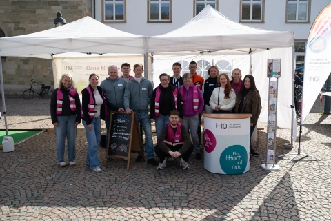 Gruppe von 13 Personen vor einem wei&szlig;en Pavillon mit Informationsstand. Einige tragen graue Jacken, andere pink-gestreifte Schals. Auf einem Aufsteller steht: &bdquo;Die! Hochschule Osnabr&uuml;ck&ldquo;. Rechts ein bedruckter Stehtisch mit Text &bdquo;Ein Hoch auf Dich.&ldquo; Kopfsteinpflaster und helle Geb&auml;ude im Hintergrund.