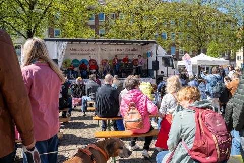 Menschen sitzen und stehen auf einem gepflasterten Platz, viele auf Holzb&auml;nken, und schauen auf eine B&uuml;hne mit wei&szlig;en Bannern, auf denen "Osna f&uuml;r alle!" steht. Auf der B&uuml;hne sitzen mehrere Personen, mikrofonierte Reden haltend. Sonnige Atmosph&auml;re mit Fr&uuml;hlingsb&auml;umen und weiteren Marktst&auml;nden im Hintergrund. Ein Hund mit braunem Fell liegt im Vordergrund links neben einer Bank.