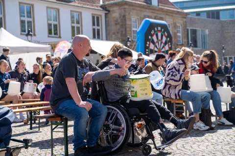 Menschen sitzen auf Holzb&auml;nken auf einem gepflasterten Platz. Im Vordergrund ein Mann mit Glatze neben einer Person im Rollstuhl, beide halten wei&szlig;e Plastikeimer. Im Hintergrund mehrere sitzende und stehende Personen, ein gro&szlig;es Dartspiel und Geb&auml;ude.