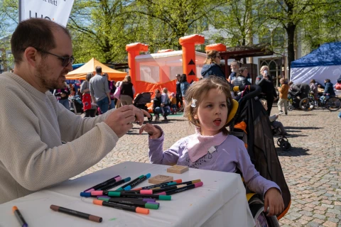 Erwachsene Person in beigem Pullover und Kind in lila Oberteil sitzen an einem kleinen wei&szlig;en Tisch auf einem gepflasterten Platz. Auf dem Tisch liegen verschiedene bunte Filzstifte. Im Hintergrund stehen Menschen vor orangefarbenen und blauen Pavillons sowie einer H&uuml;pfburg in leuchtendem Orange. Gr&uuml;n belaubte B&auml;ume s&auml;umen den Platz.