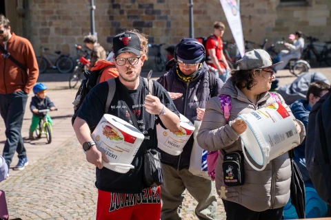 Personen in legerer Kleidung mit gro&szlig;en wei&szlig;en Plastikeimern, auf denen bunte Logos und Texte zu sehen sind, offenbar in Bewegung. Im Hintergrund mehrere Personen sowie Fahrr&auml;der und ein Geb&auml;ude mit Steinmauer. Sonniges Tageslicht.