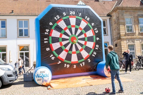 Aufblasbare Dartscheibe in Schwarz, Wei&szlig;, Gr&uuml;n und Rot mit gro&szlig;en Zahlen von 1 bis 20. Eine Person mit blauer Jacke h&auml;lt einen Fu&szlig;ball und steht vor der Scheibe. Ein weiterer Fu&szlig;ball liegt am Boden. Links ein wei&szlig;er Transporter, rechts Fahrr&auml;der, im Hintergrund eine Hausfassade mit Fenstern. Auf dem linken Ball der Schriftzug: &bdquo;Dein Verein bist du!&ldquo;.