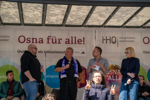 Vier Personen stehen auf einer B&uuml;hne vor einem bedruckten Banner mit dem Text &bdquo;Osna f&uuml;r alle!&ldquo;. Zwei M&auml;nner halten Mikrofone, w&auml;hrend sie miteinander sprechen. Links und rechts von ihnen stehen eine Frau und ein Mann, teils mit Mikrofon in der Hand. Vor der B&uuml;hne hebt ein Kind in einem blauen Oberteil beide H&auml;nde und formt mit den Fingern ein Friedenszeichen. Im Hintergrund sind bunte Kreise und Logos auf dem Banner erkennbar, darunter &bdquo;HHO &ndash; wir gestalten miteinander&ldquo;.