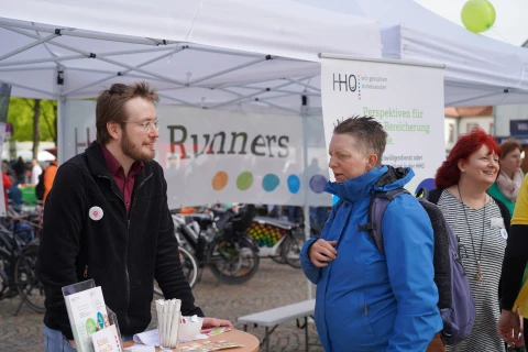 Ein Stand mit wei&szlig;em Pavillon, auf dem Flyer und ein Aufsteller mit der Aufschrift &bdquo;Sch&ouml;n, dass Du&hellip;&ldquo; liegen. Links ein Banner mit &bdquo;Runners&ldquo; und farbigen Punkten, rechts ein Roll-Up mit Text: &bdquo;HHO &ndash; Perspektiven f&uuml;r Arbeits-/Bereicherung&ldquo;. Im Hintergrund Fahrr&auml;der, Ballons und weitere Menschen.