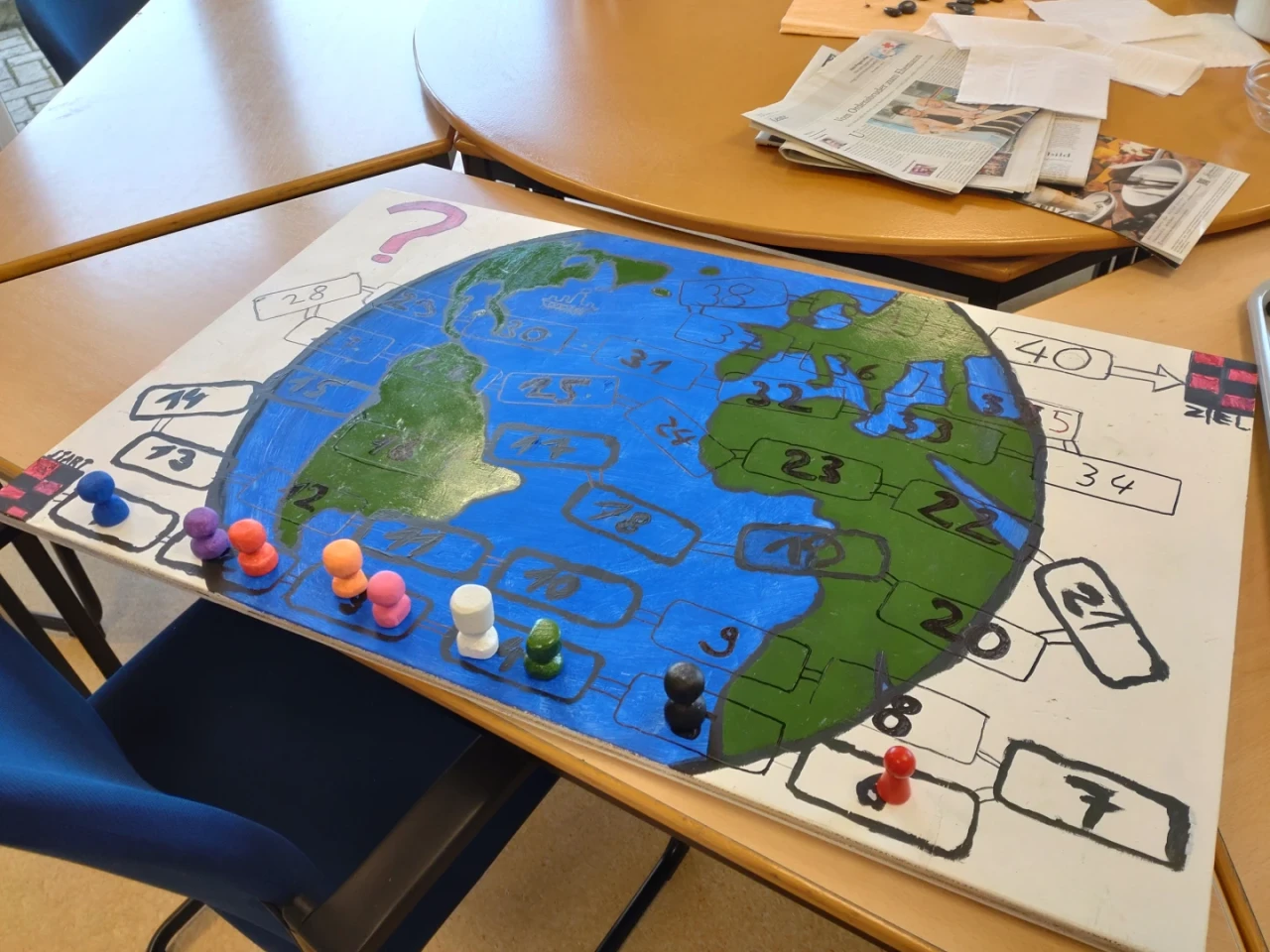 Brettspiel auf Tisch mit gemaltem Weltkarten-Design: Ozeane blau, Kontinente gr&uuml;n, nummerierte Felder auf der Karte und am Rand. Acht farbige Spielfiguren (u. a. orange, pink, schwarz, wei&szlig;) an der Startposition. Im Hintergrund Holz- und Metalltische mit Stapel Papier und Zeitungen.