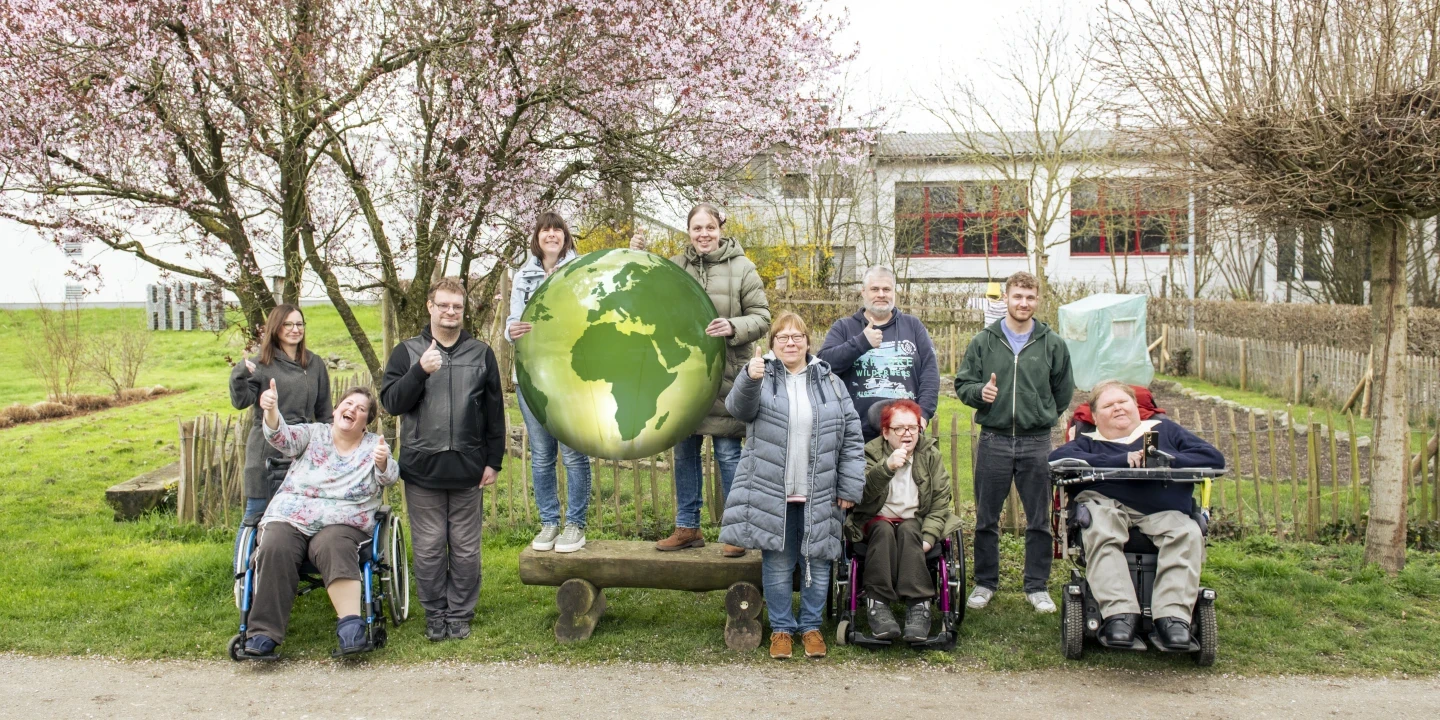 Eine Gruppe von zehn Personen, darunter Rollstuhlfahrer, steht und sitzt auf einer Gr&uuml;nfl&auml;che vor einem bl&uuml;henden Baum und einem Geb&auml;ude mit roten Fenstern. Sie halten einen gro&szlig;en Globus mit der Oberfl&auml;che Afrikas und Europas in der Mitte. Alle l&auml;cheln und zeigen Daumen nach oben.