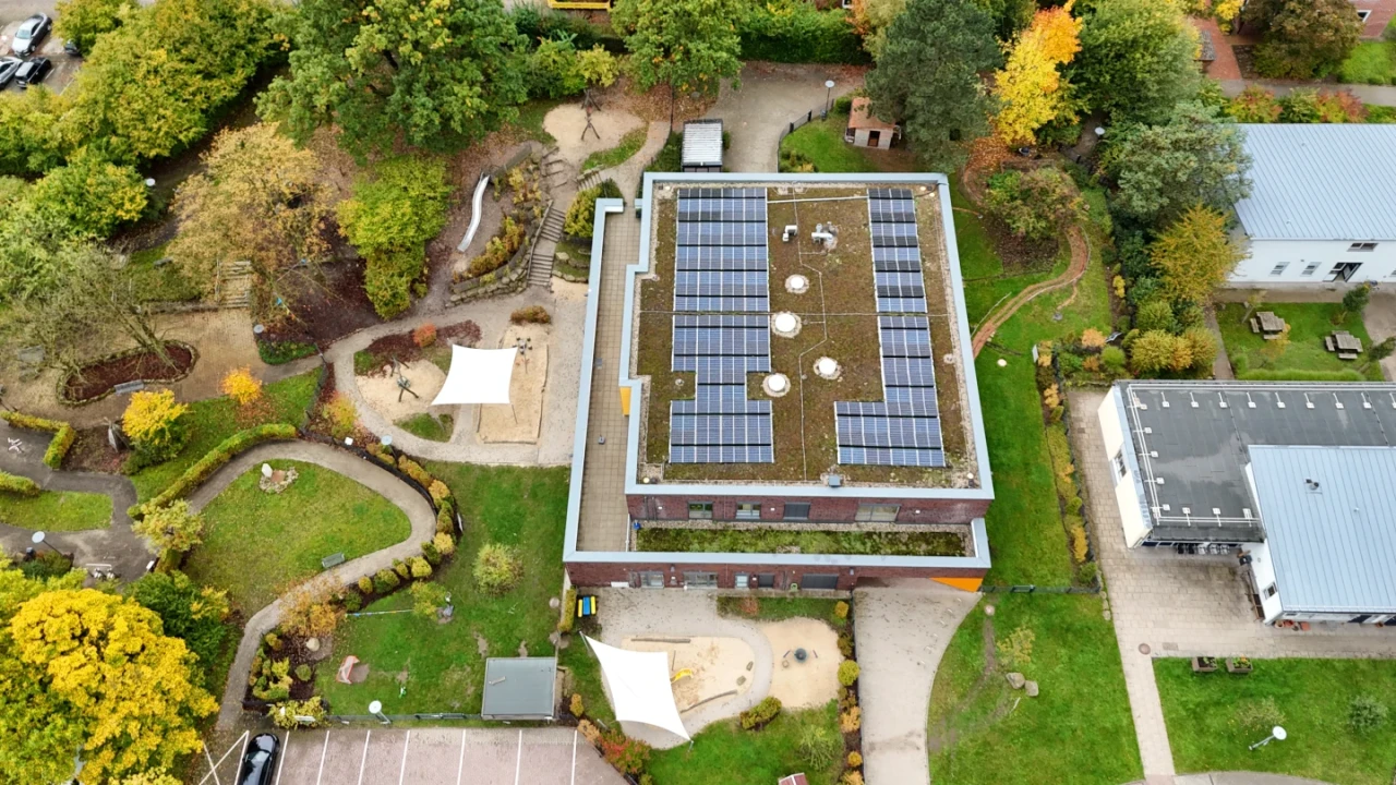 Flachdachgeb&auml;ude mit Solarpanelen und begr&uuml;ntem Dach, umgeben von Gr&uuml;nanlagen, B&auml;umen und geschwungenen Wegen. Links ein Spielplatzbereich mit Sandfl&auml;chen und Sonnensegeln, unten ein Parkplatz mit Autos, rechts ein weiteres Geb&auml;ude mit grauem Dach.