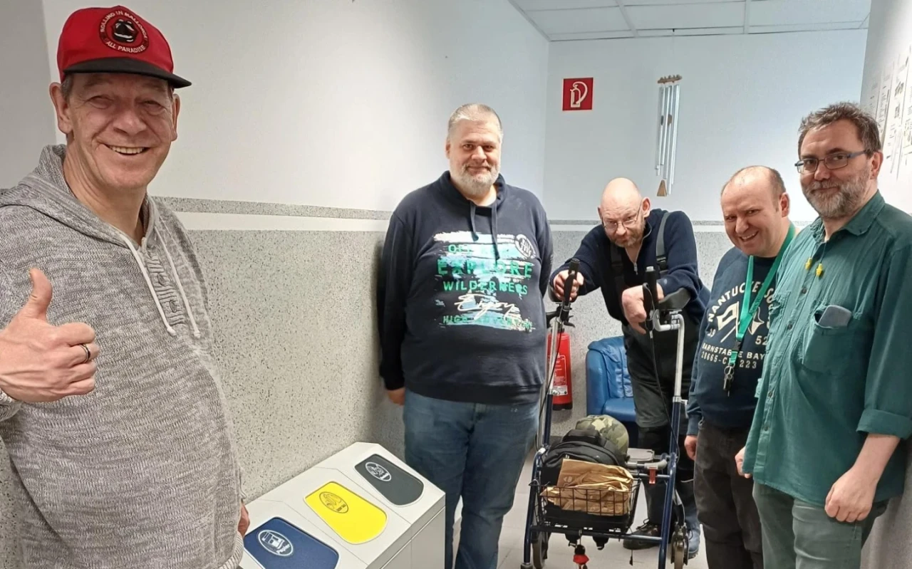 F&uuml;nf M&auml;nner in einem Flur mit wei&szlig;en W&auml;nden und grauem Boden, drei stehend, einer mit Daumen nach oben und ein anderer auf einen Rollator gest&uuml;tzt. Links zwei Abfallbeh&auml;lter mit gelbem und blauem Deckel, rechts ein Feuerl&ouml;scher an der Wand. Deckenbeleuchtung und Notausgangsschild erkennbar.