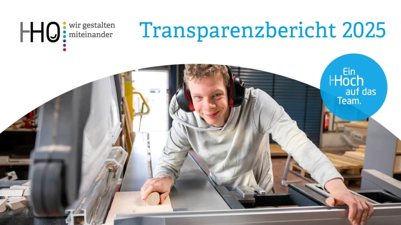 Mann mit Geh&ouml;rschutz und grauem Pullover lehnt sich an eine gro&szlig;e Tischs&auml;ge in einer Werkstatt, l&auml;chelnd. Im oberen Bereich ein gebogener Text: "HHO - wir gestalten miteinander. Transparenzbericht 2025". Ein blauer Kreis rechts enth&auml;lt die Worte "Ein HHOch auf das Team".