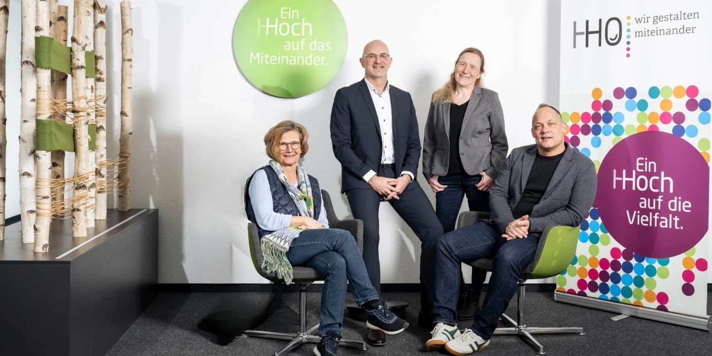 Vier Personen in einem B&uuml;robereich. Zwei sitzen auf gr&uuml;nen St&uuml;hlen, zwei stehen dahinter. Links birkenartige Dekoration, rechts zwei runde Schilder: "Ein Hoch auf das Miteinander" (gr&uuml;n) und "Ein Hoch auf die Vielfalt" (bunt). Hinten "HHO &ndash; wir gestalten miteinander"-Logo auf wei&szlig;em Hintergrund.