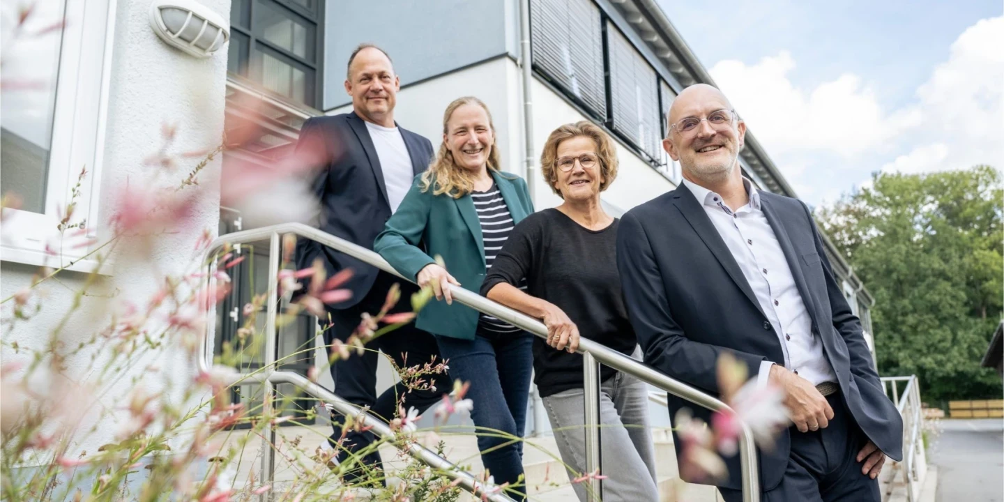 Vier Personen stehen nebeneinander auf einer Treppe mit Metallgel&auml;nder vor einem Geb&auml;ude mit wei&szlig;en und grauen Fassaden und gro&szlig;en Fenstern. Im Vordergrund unscharfe Blumen. Im Hintergrund gr&uuml;ne B&auml;ume und blauer Himmel mit wei&szlig;en Wolken.