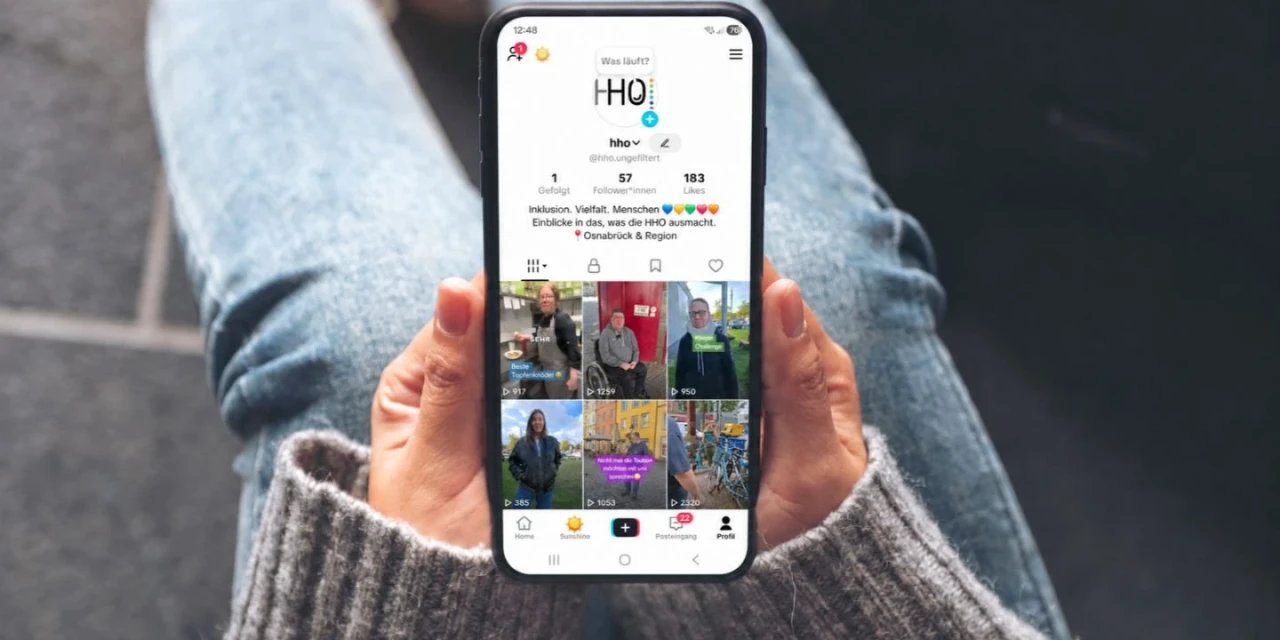 Smartphone in beiden H&auml;nden gehalten, auf dem ein ge&ouml;ffneter TikTok-Profilbildschirm sichtbar ist. Der Bildschirm zeigt das Profil "hho", mit Profilbild, 57 Abonnenten, 153 Likes und mehreren Videovorschauen in einem 3x3-Raster. Person tr&auml;gt eine graue, gestrickte Handbekleidung, Hintergrund unscharf, blau-graue Jeans sichtbar.