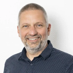 Mittelalte Person mit kurzem, grauem Haar und Bart, tr&auml;gt ein dunkelblaues, fein gepunktetes Hemd. Die Person l&auml;chelt, der Hintergrund ist eine wei&szlig;e, strukturierte Wand.