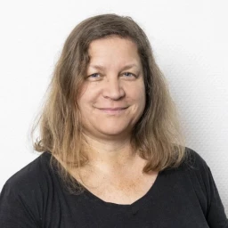 Frau mit schulterlangem, welligem, mittelbraunem Haar und schwarzen Oberteil vor wei&szlig;em Hintergrund mit leicht strukturiertem Muster. Gesicht leicht l&auml;chelnd, direkt zur Kamera gewandt.