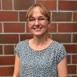 Frau mit blonden Haaren und einer Brille, l&auml;chelt in die Kamera. Sie tr&auml;gt ein blaues Oberteil mit wei&szlig;em Blumenmuster. Der Hintergrund besteht aus einer Ziegelwand mit r&ouml;tlich-braunen und grauen Steinen.