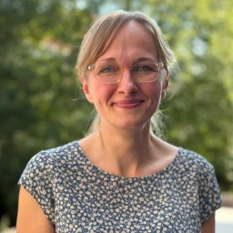 Frau mit schulterlangem, blondem Haar, das seitlich gescheitelt ist, und einer runden Brille. Sie tr&auml;gt ein kurz&auml;rmeliges, blaues Kleid mit kleinen wei&szlig;en Blumenmustern. Im Hintergrund gr&uuml;ne, verschwommene Vegetation bei Tageslicht.