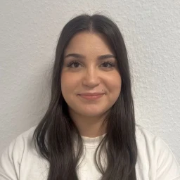 Portr&auml;t einer Person mit langen, glatten, dunklen Haaren, die &uuml;ber die Schultern fallen. Die Person tr&auml;gt ein wei&szlig;es Oberteil und blickt direkt in die Kamera. Der Hintergrund ist eine wei&szlig;e, strukturierte Wand.