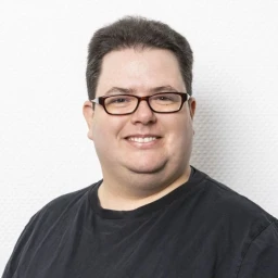 Mann mittleren Alters mit kurzen, dunklen Haaren und Brille, l&auml;chelnd, in schwarzem T-Shirt gekleidet. Hintergrund aus wei&szlig;er, strukturierter Wand.