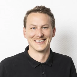 Mann mit hellem Teint, kurzem blondem Haar und Schnurrbart, tr&auml;gt ein schwarzes Poloshirt und l&auml;chelt, vor einer wei&szlig;en, strukturierten Wand.