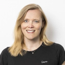 Portr&auml;t einer l&auml;chelnden Frau mit schulterlangem blondem Haar, blau-grauen Augen und hellem Teint. Sie tr&auml;gt ein schwarzes T-Shirt mit dem Schriftzug &bdquo;love&ldquo; in wei&szlig;er Schreibschrift auf der Brust. Hintergrund ist eine wei&szlig;e, leicht strukturierte Fl&auml;che.