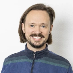 Mann mit schulterlangem braunem Haar, Bart und breitem L&auml;cheln, tr&auml;gt einen blauen Pullover mit horizontalen dunkelblauen und gr&uuml;nen Streifen, vor einer wei&szlig;en strukturierten Wand.