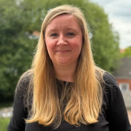 Frau mit langen blonden Haaren und schwarzem Oberteil, l&auml;chelnd in die Kamera blickend, im Freien aufgenommen. Im Hintergrund gr&uuml;ne B&auml;ume, ein grau bew&ouml;lkter Himmel und ein Teil eines Ziegelhauses.