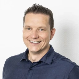 Mittelgro&szlig;er Mann mit kurzem, braunem Haar, das nach hinten gestylt ist. Er tr&auml;gt ein dunkelblaues Hemd, blickt l&auml;chelnd nach vorne und steht vor einer wei&szlig;en, strukturierten Wand.