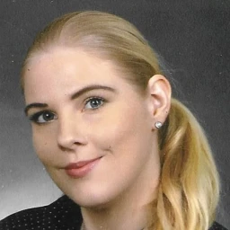 Portr&auml;t einer l&auml;chelnden Person mit blonden, zu einem Pferdeschwanz gebundenen Haaren. Sie tr&auml;gt ein schwarzes Hemd mit wei&szlig;en Punkten und eine silberne Halskette. Der Hintergrund ist grau.