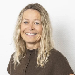 Frau mit langen, welligen blonden Haaren, brauner Knopfbluse, l&auml;chelnd, vor einer wei&szlig;en strukturierten Wand.