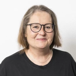 Frau mit schulterlangem braunem Haar, schwarzer Brille und schwarzem Oberteil vor einer wei&szlig;en, leicht strukturierten Wand, l&auml;chelnd in die Kamera blickend.