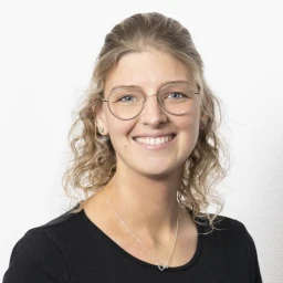 Oberk&ouml;rper einer Person mit hellbraunen, lockigen Haaren, die nach hinten gebunden sind. Schwarzes Oberteil mit Rundhalsausschnitt, eine schlichte silberne Halskette und kleine silberne Ohrringe. Hintergrund in einfarbigem Wei&szlig;.