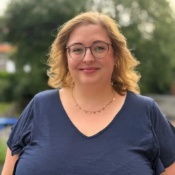 Frau mit schulterlangem, welligem blondem Haar und Brille, blaue Bluse mit V-Ausschnitt, goldene Halskette mit kleinen Anh&auml;ngern. Hintergrund unscharf mit gr&uuml;nen B&auml;umen und Gartenbereich.