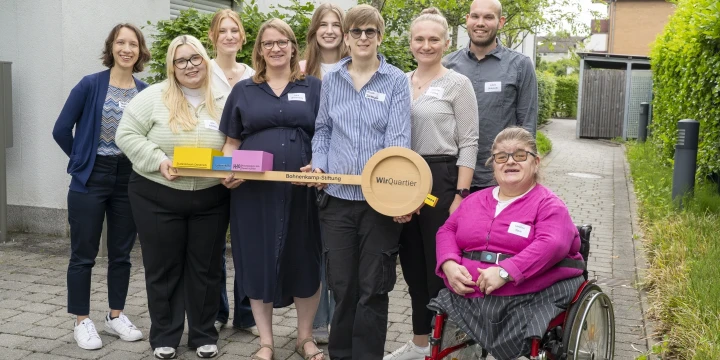 Eine Gruppe von neun Personen steht auf einem gepflasterten Weg vor einem Geb&auml;ude mit wei&szlig;en W&auml;nden. Sie halten gemeinsam eine gro&szlig;e Holzattrappe eines Schl&uuml;ssels mit der Aufschrift &bdquo;WirQuartier&ldquo;. Eine Person sitzt im Rollstuhl, tr&auml;gt ein pinkfarbenes Oberteil und h&auml;lt den Schl&uuml;ssel. Im Hintergrund sind gr&uuml;ne B&auml;ume und weitere H&auml;user erkennbar.