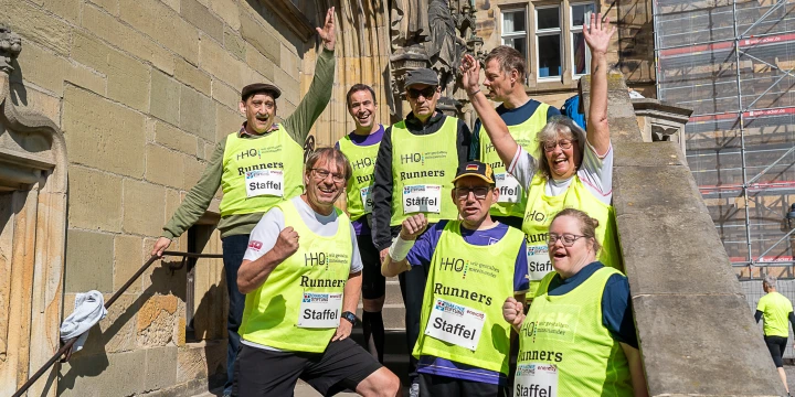 Gruppe von Personen in gelben &auml;rmellosen Laufwesten mit der Aufschrift "HQ Runners Staffel" auf einer Steintreppe vor einem historischen Geb&auml;ude, teils mit erhobenen Armen. Rechts im Hintergrund moderne Ger&uuml;ststruktur, links historisches Mauerwerk. Sonnenlicht erhellt die Szene.