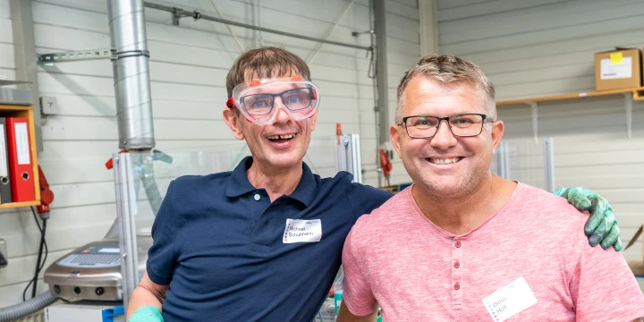 Zwei Personen in Arbeitsumgebung, links im dunkelblauen Poloshirt mit gr&uuml;nen Handschuhen und rechts im rosa T-Shirt mit Namensschild. Hintergrund: industrielle Ger&auml;te, beige Wand, Regale mit Kartons.