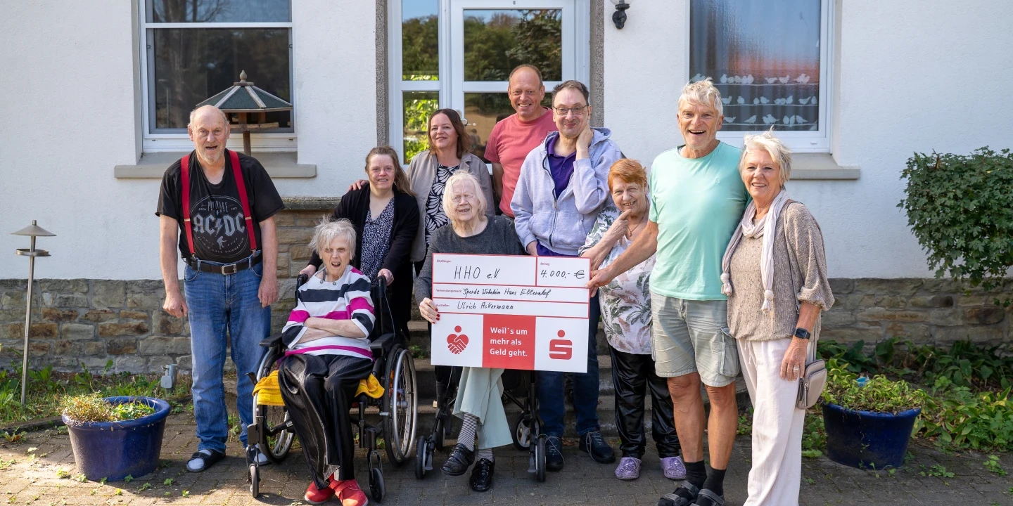 Gruppe von zehn Personen vor einem wei&szlig;en Geb&auml;ude mit der Aufschrift "Ellenhof". In der Mitte halten zwei Personen einen gro&szlig;en symbolischen Scheck mit der Aufschrift "HHO e. V." und einer Summe von 900 &euro;. Eine Person sitzt im Rollstuhl, umgeben von weiteren Personen, die l&auml;cheln. Zwei blaue Pflanzent&ouml;pfe stehen links und rechts des Eingangs.