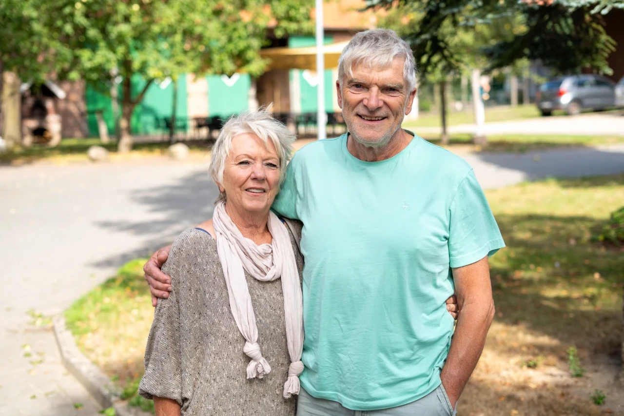 &Auml;ltere Frau mit hellem, schulterlangem Haar tr&auml;gt einen hellgrauen Pullover und einen beigefarbenen Schal, steht Arm in Arm mit einem Mann mit grauen Haaren in einem hellgr&uuml;nen T-Shirt. Im Hintergrund Gehweg, Wiese, B&auml;ume und ein Geb&auml;ude mit gr&uuml;ner Fassade.