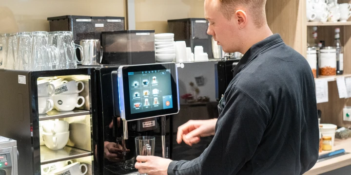Person mit schwarzem Oberteil bedient eine schwarze Kaffeevollautomat-Maschine mit Touchscreen; daneben Regale mit wei&szlig;en Tassen, Gl&auml;sern, Kaffeebeh&auml;ltern und Utensilien. Glasschrank mit weiteren Gl&auml;sern im Hintergrund.