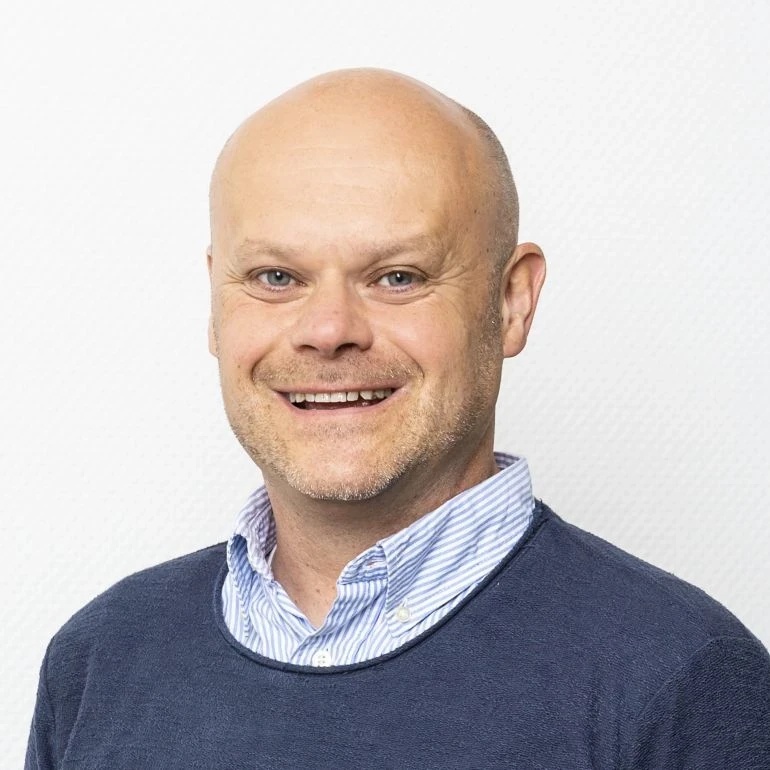 Portr&auml;t eines l&auml;chelnden Mannes mit Glatze, hellem Teint und braunen Augen. Er tr&auml;gt ein hellblau-wei&szlig; gestreiftes Hemd unter einem dunkelblauen Pullover. Der Hintergrund ist eine glatte, wei&szlig;e Wand.