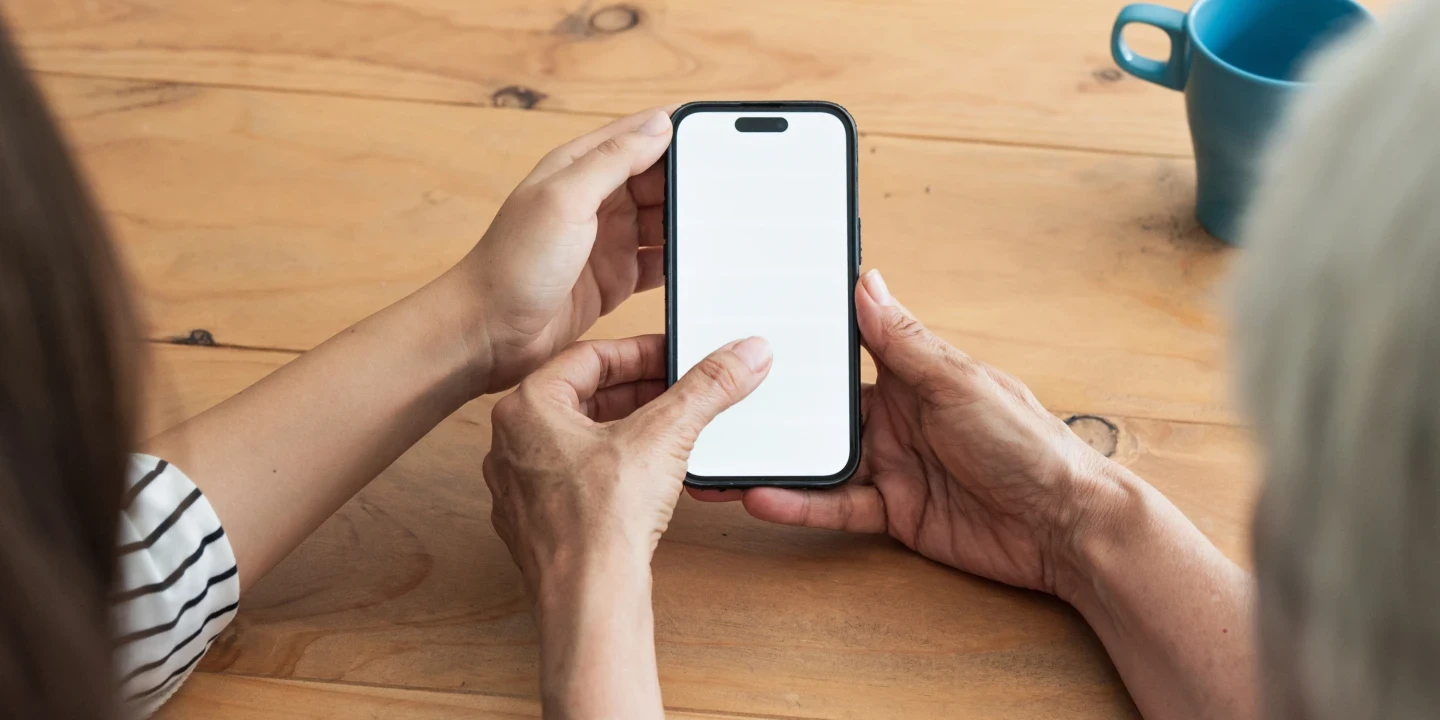 Zwei H&auml;nde halten ein Smartphone mit wei&szlig;em, leerem Bildschirm &uuml;ber einem Holztisch. Links daneben sitzt eine Person mit gestreiftem Oberteil, rechts eine Person mit grauen Haaren. Im Hintergrund eine blaue Tasse.