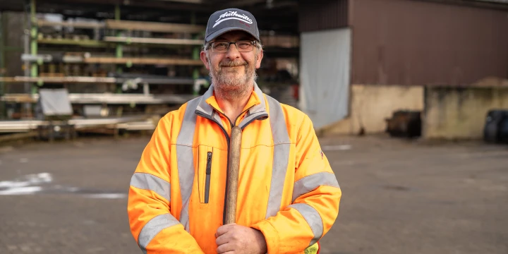 Person in einer auff&auml;lligen orangefarbenen Warnschutzjacke mit reflektierenden Streifen h&auml;lt ein Werkzeugstiel vor sich. Im Hintergrund ein Hof mit Kopfsteinpflaster, Regale mit Holzmaterialien und ein braunes Geb&auml;ude mit einem ge&ouml;ffneten Tor. Rechts einige schwarze Gegenst&auml;nde.
