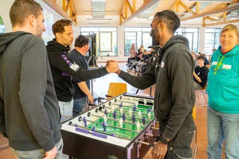 Vier Personen an einem Tischkicker, zwei davon sch&uuml;tteln einander die H&auml;nde. Der Tischkicker hat gr&uuml;ne Spielfl&auml;che und violette Spielfiguren. Im Hintergrund ein heller Raum mit Holzdecke, mehreren Fenstern, St&uuml;hlen und sitzenden Personen.
