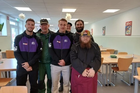 Gruppe von sechs Personen, teils in lila-schwarzen Trainingsjacken mit Logoaufdruck, eine Person in gr&uuml;ner Uniform und eine in dunkler Kleidung mit langem Bart und roter Sch&uuml;rze. Alle stehen in einem Speisesaal mit leeren Holztischen und St&uuml;hlen. An den W&auml;nden h&auml;ngen bunte Poster, an der Decke befinden sich wei&szlig;e Sterne als Dekoration.