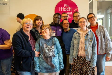 Gruppe von neun Menschen unterschiedlicher Kleidung und Hautfarbe, einige in Freizeitkleidung, ein M&auml;dchen mit Pullover mit Katzenmotiv. Hintergrund zeigt eine wei&szlig;e Wand mit einem pinken Kreis und Schriftzug "Ein Hoch auf dich." sowie einen gelben Kreis.