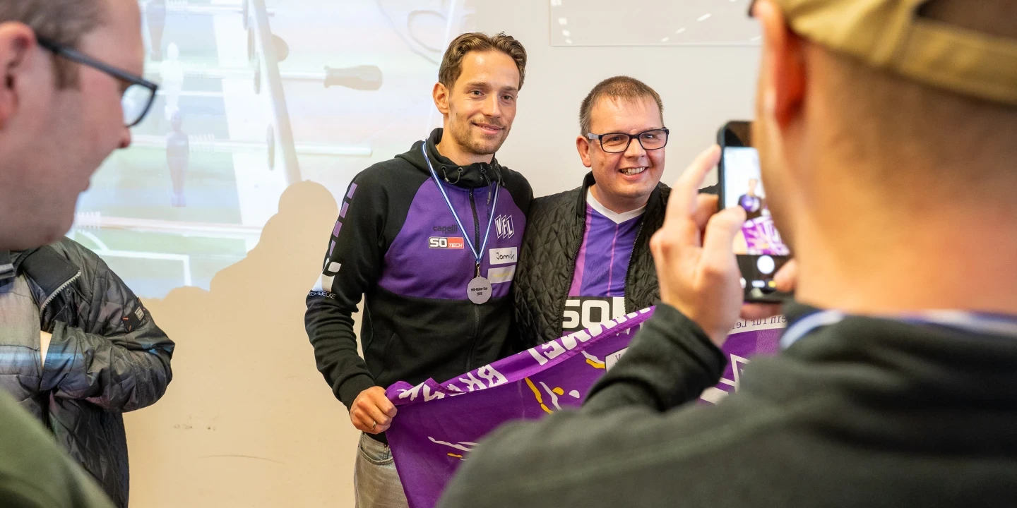 Zwei Personen in Kleidung mit violetten und wei&szlig;en Akzenten halten ein violettes Banner mit Schrift. Im Vordergrund fotografiert eine Person mit beigefarbener Kappe die Szene. Hinten links ein Tischkicker und eine Projektion, rechts ein wei&szlig;es Schild mit schwarzem Text.