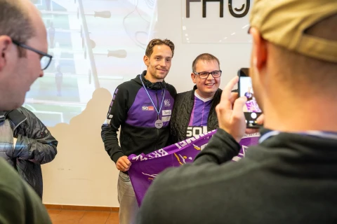Zwei Personen in Kleidung mit violetten und wei&szlig;en Akzenten halten ein violettes Banner mit Schrift. Im Vordergrund fotografiert eine Person mit beigefarbener Kappe die Szene. Hinten links ein Tischkicker und eine Projektion, rechts ein wei&szlig;es Schild mit schwarzem Text.